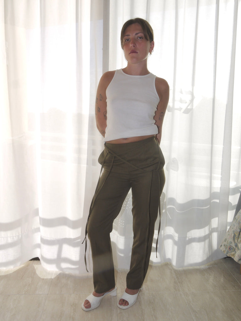 Model wearing a long-sleeve black top with asymmetrical seams, paired with iconic olive green Maison Margiela pants featuring deconstructed strap detailing hanging from the waistband. Styled with black-and-white printed heels for a conceptual, avant-garde look. Shot against a plain white background.