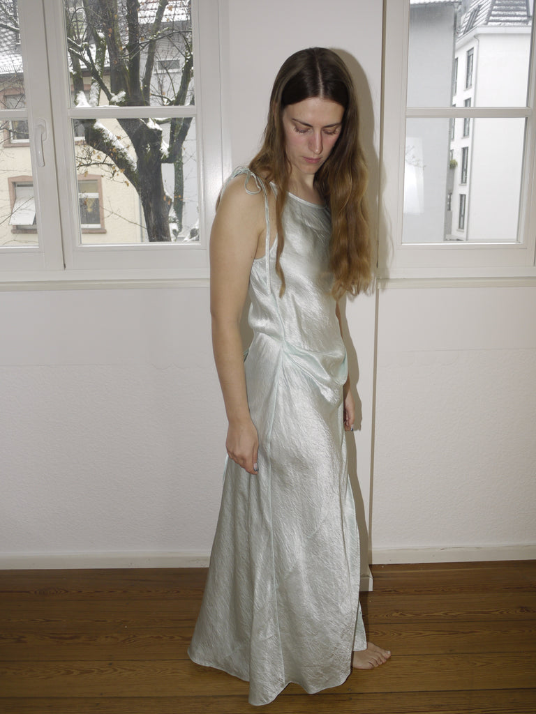 Light sage green satin slip dress by Acne Studios, fluid silhouette with subtle shimmer, minimalist eveningwear aesthetic, modeled indoors in natural daylight.