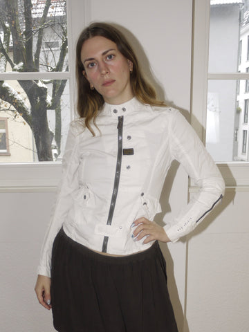 Original G-Star white fitted denim jacket with black hardware and asymmetric button details, structured silhouette with a modern military edge, styled with pleated mini skirt, modeled indoors.