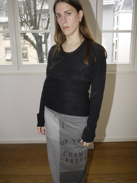 Avant-garde black long sleeve top with layered textures, inspired by Dries Van Noten’s signature deconstructed aesthetic, styled with grey Y2K print skirt, worn by model in natural light.