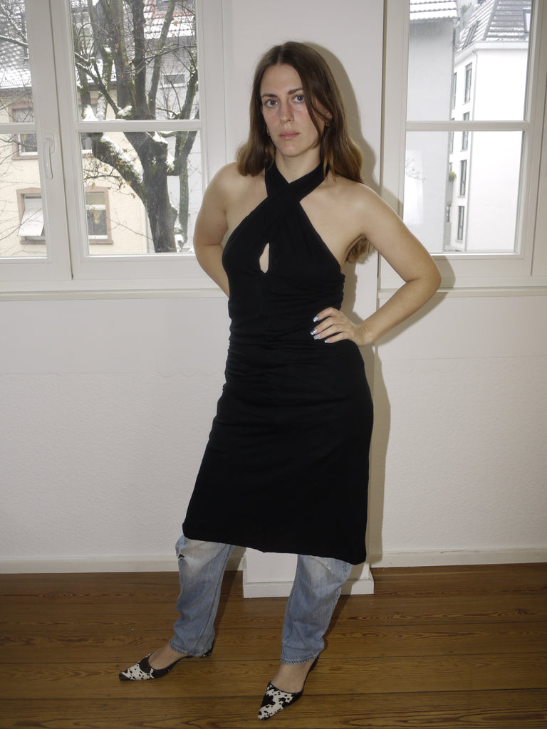 Vintage black Margiela-style halterneck dress with beige side stripe and open back, styled over jeans and modeled indoors in natural light. Draped fit with a modern, deconstructed vibe reminiscent of Maison Margiela aesthetics.