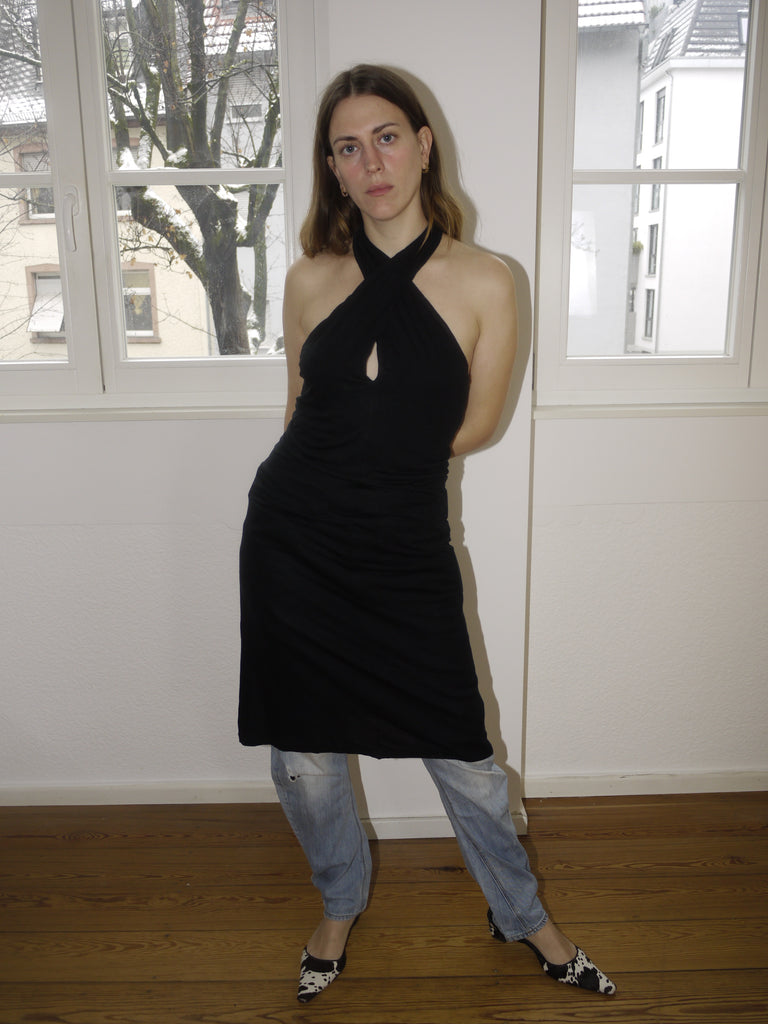 Vintage black Margiela-style halterneck dress with beige side stripe and open back, styled over jeans and modeled indoors in natural light. Draped fit with a modern, deconstructed vibe reminiscent of Maison Margiela aesthetics.