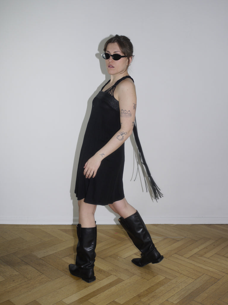 Y2K Cop Copine black strappy mini dress with asymmetrical fringe detail on the side. Styled with knee-high black leather boots and narrow sunglasses for a bold early 2000s look, inspired by avant-garde Parisian streetwear.