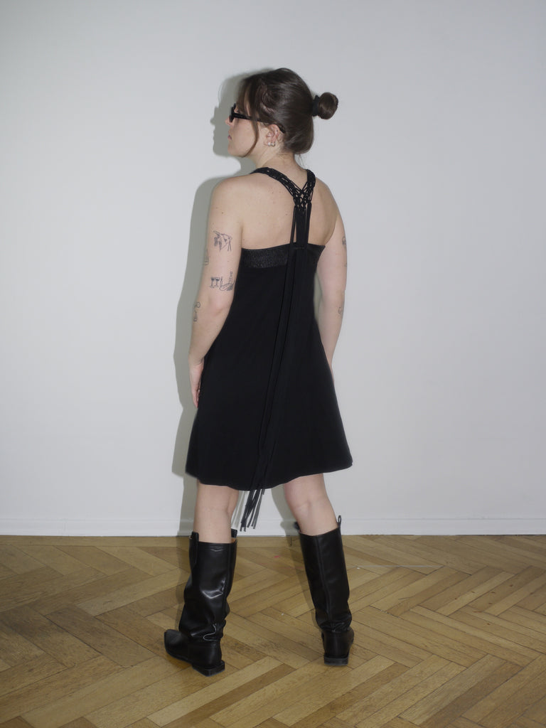 Y2K Cop Copine black strappy mini dress with asymmetrical fringe detail on the side. Styled with knee-high black leather boots and narrow sunglasses for a bold early 2000s look, inspired by avant-garde Parisian streetwear.