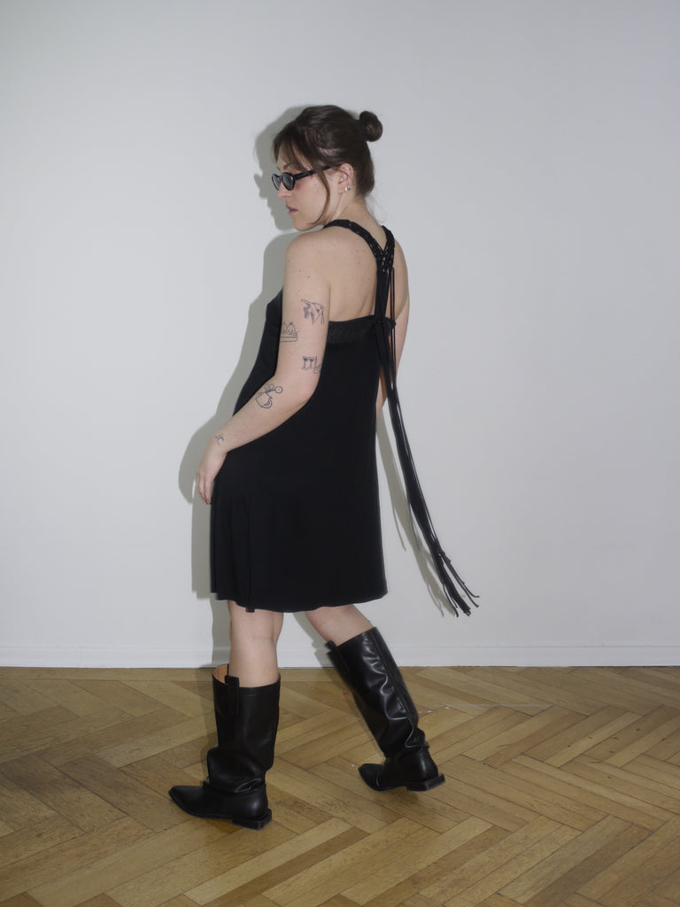 Y2K Cop Copine black strappy mini dress with asymmetrical fringe detail on the side. Styled with knee-high black leather boots and narrow sunglasses for a bold early 2000s look, inspired by avant-garde Parisian streetwear.