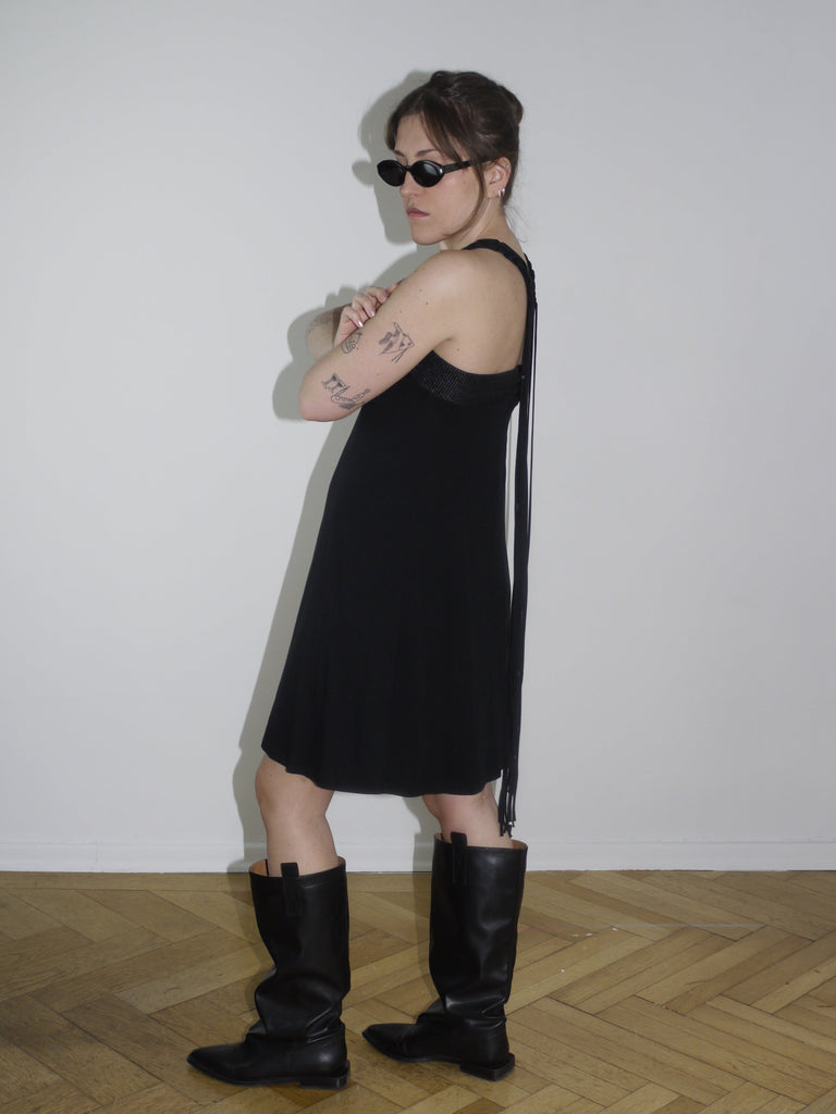 Y2K Cop Copine black strappy mini dress with asymmetrical fringe detail on the side. Styled with knee-high black leather boots and narrow sunglasses for a bold early 2000s look, inspired by avant-garde Parisian streetwear.