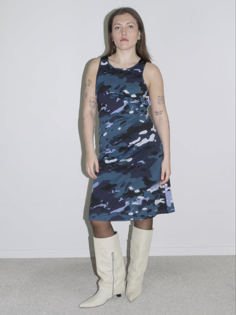 A woman wearing a sleeveless blue and black camouflage print dress paired with white knee-high boots. The look blends a bold, sporty aesthetic with a structured silhouette, inspired by streetwear and utilitarian styles.