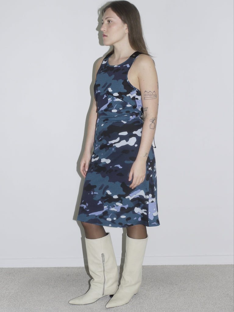 A woman wearing a sleeveless blue and black camouflage print dress paired with white knee-high boots. The look blends a bold, sporty aesthetic with a structured silhouette, inspired by streetwear and utilitarian styles.