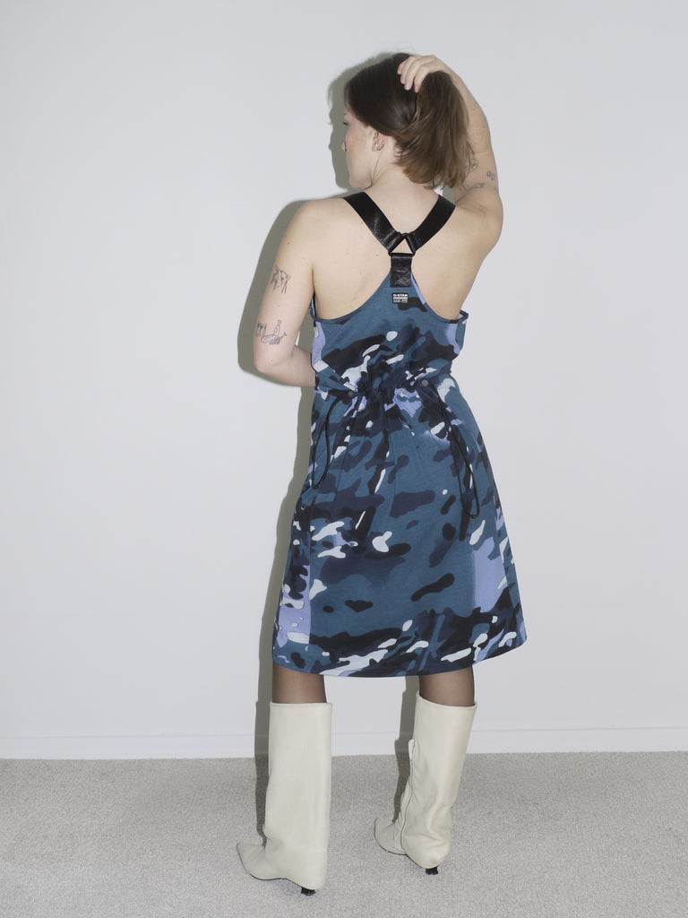 A woman wearing a sleeveless blue and black camouflage print dress paired with white knee-high boots. The look blends a bold, sporty aesthetic with a structured silhouette, inspired by streetwear and utilitarian styles.