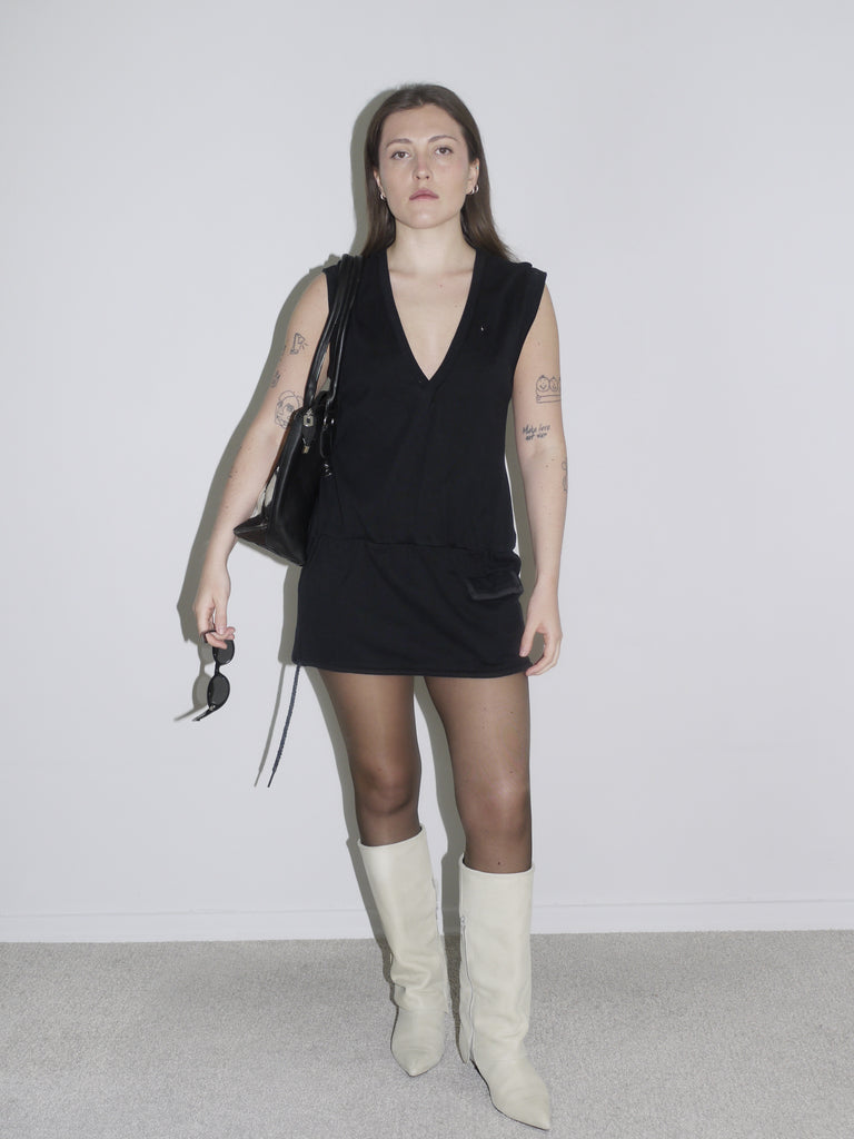 A minimal black V-neck mini dress styled with white high-heeled boots and a black shoulder bag. The outfit has a sleek, edgy feel, reminiscent of Helmut Lang or early 2000s Givenchy vibes.