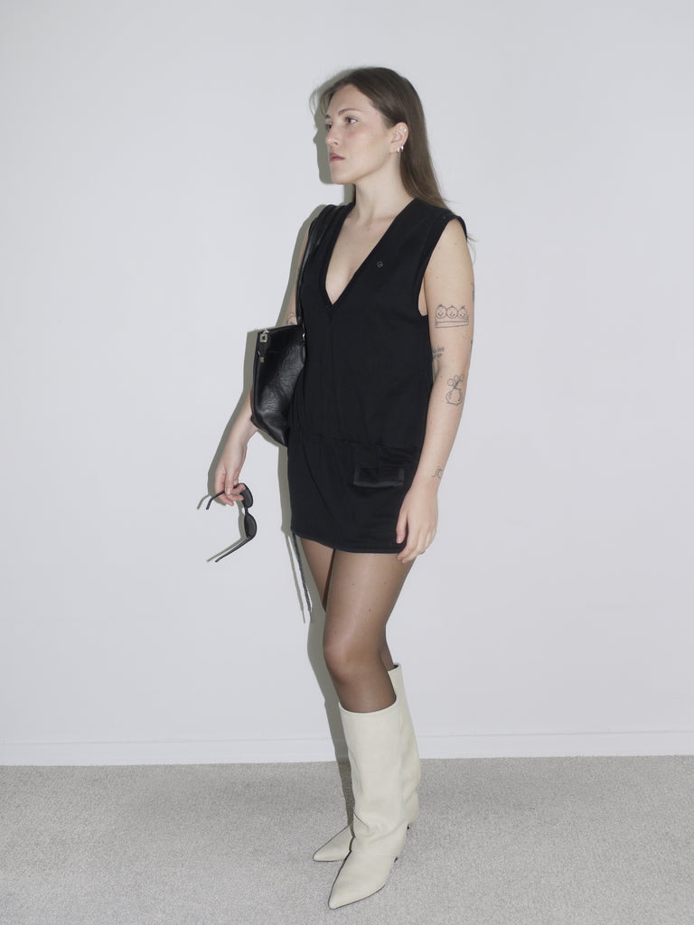 A minimal black V-neck mini dress styled with white high-heeled boots and a black shoulder bag. The outfit has a sleek, edgy feel, reminiscent of Helmut Lang or early 2000s Givenchy vibes.