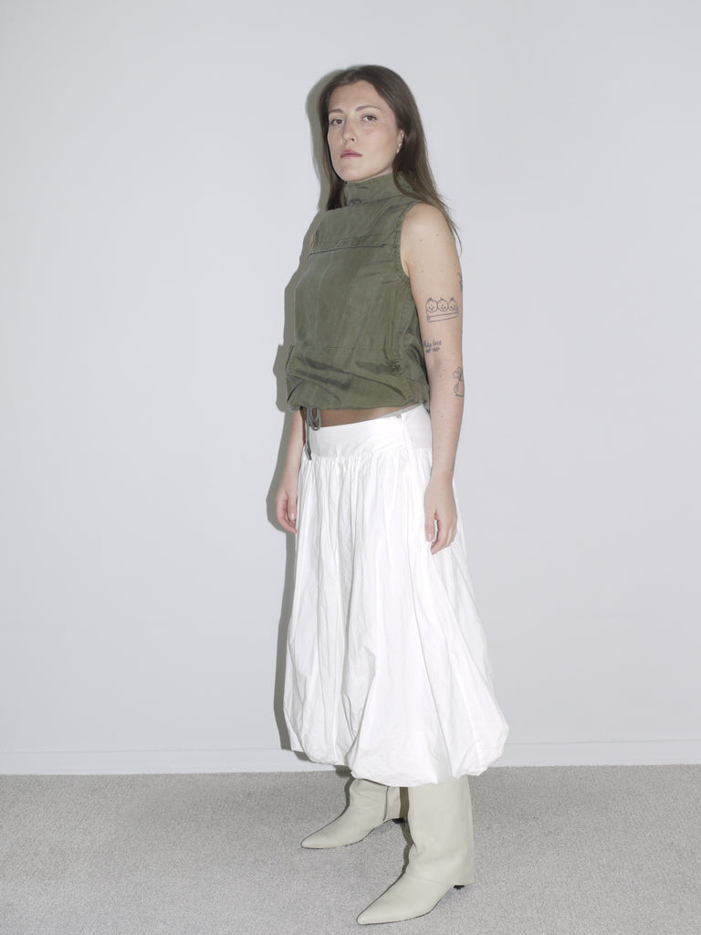 Minimalist desert-inspired outfit with an olive green sleeveless high-neck top featuring drawstring and flap pockets, styled with a voluminous white balloon skirt and cream western boots.