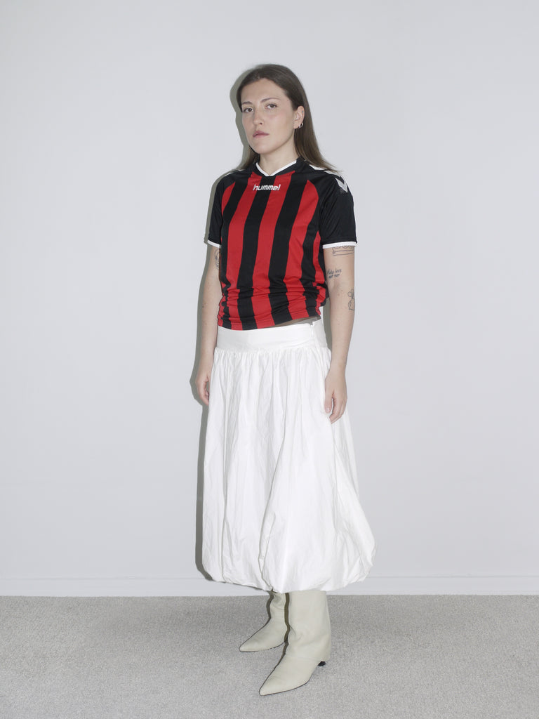 Model wearing a vintage Hummel football jersey in red and black stripes, styled with a voluminous white balloon skirt and off-white cowboy boots. A sporty-feminine mix with early 2000s energy — like AC Milan meets Paloma Wool.