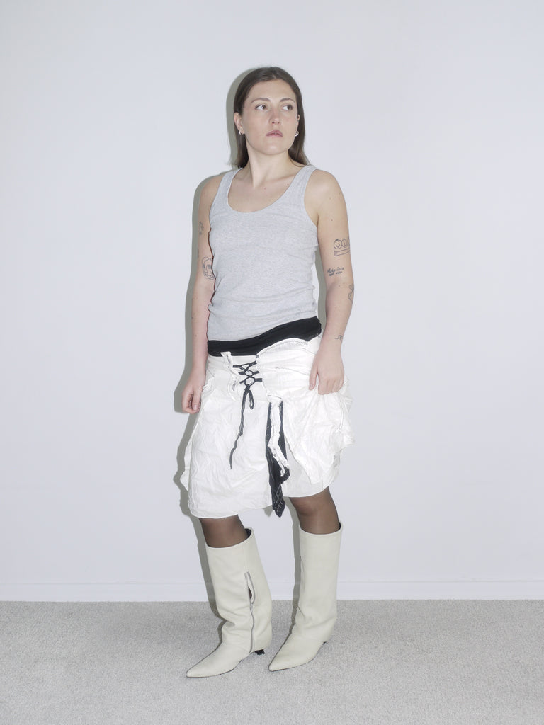 Model wearing a crinkled white parachute mini skirt with lace-up detail over black lining, paired with a grey ribbed tank top and off-white cowboy boots. A bold Y2K layering look with techwear and utility vibes — inspired by Diesel and early G-Star.