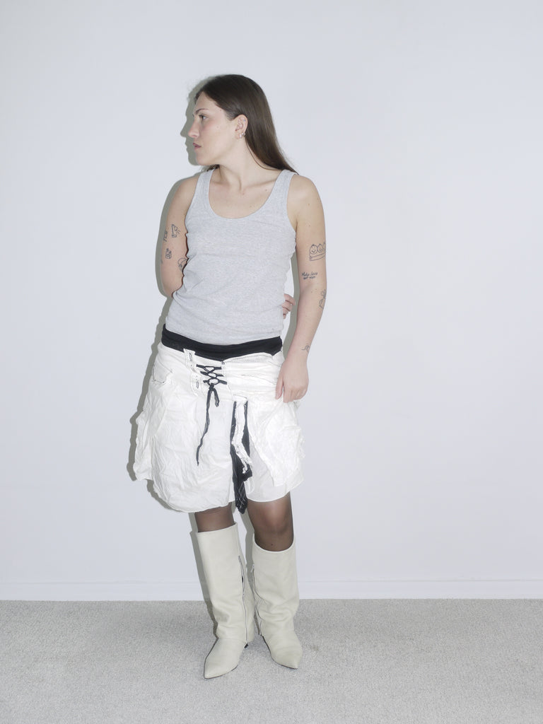 Model wearing a crinkled white parachute mini skirt with lace-up detail over black lining, paired with a grey ribbed tank top and off-white cowboy boots. A bold Y2K layering look with techwear and utility vibes — inspired by Diesel and early G-Star.