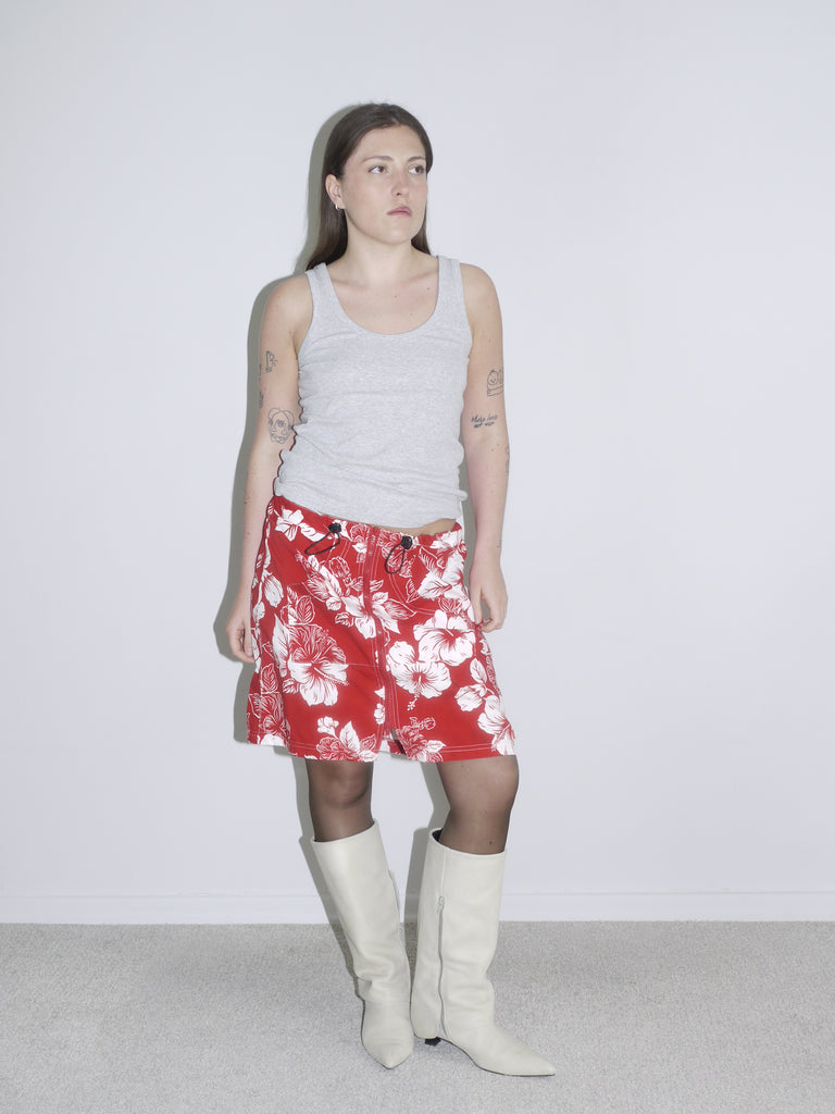 Side view of a red and white hibiscus print drawstring skirt styled with a grey tank top and off-white cowboy boots. Vintage surf aesthetic meets Y2K Western twist — think early 2000s beachcore with a fashion edge.