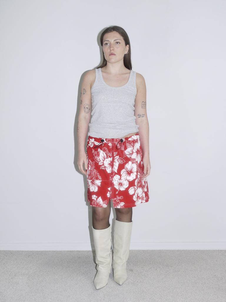 Side view of a red and white hibiscus print drawstring skirt styled with a grey tank top and off-white cowboy boots. Vintage surf aesthetic meets Y2K Western twist — think early 2000s beachcore with a fashion edge.