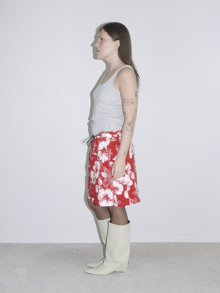 Side view of a red and white hibiscus print drawstring skirt styled with a grey tank top and off-white cowboy boots. Vintage surf aesthetic meets Y2K Western twist — think early 2000s beachcore with a fashion edge.