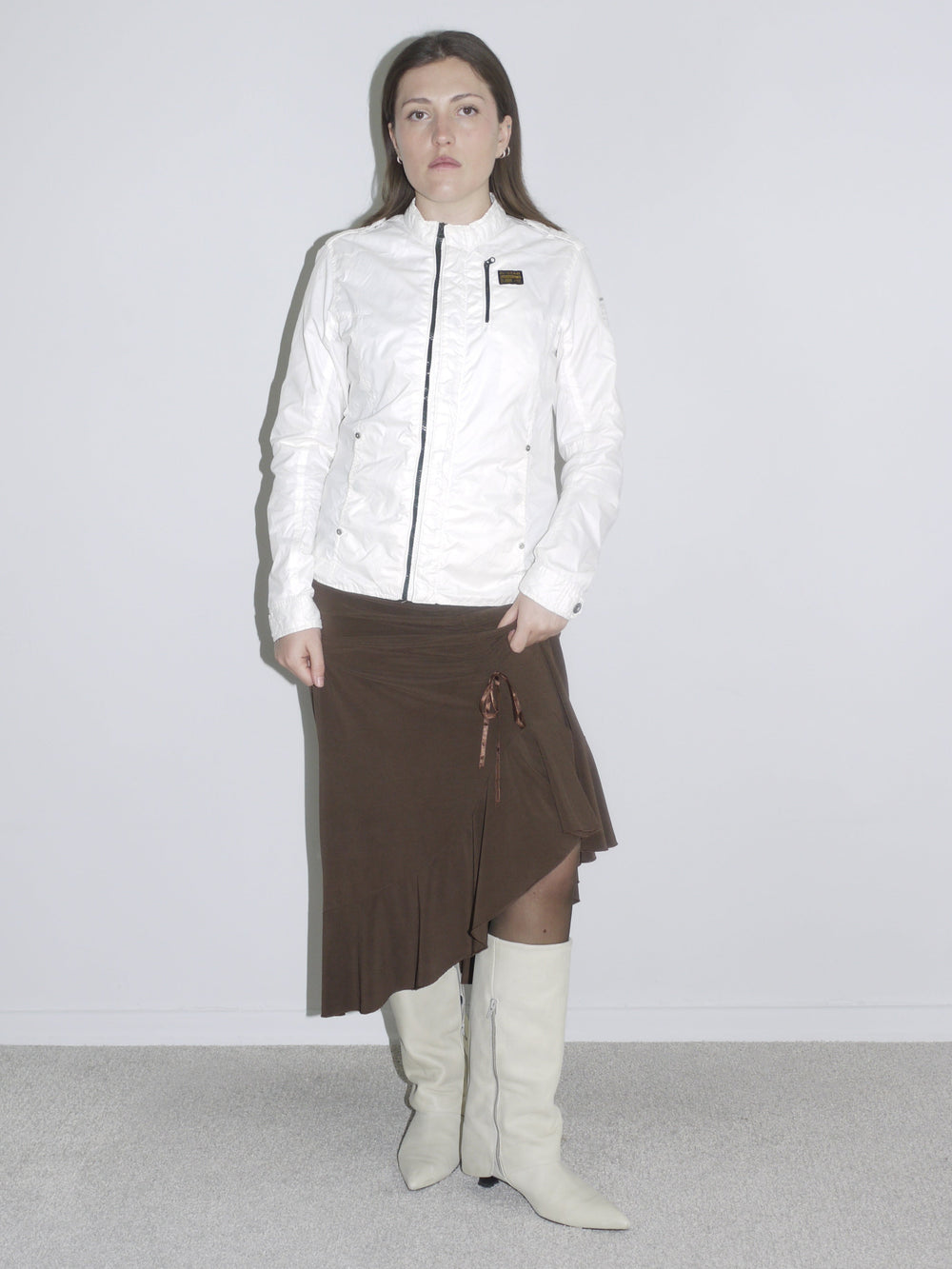 Model wearing a fitted white zip-up jacket with logo patch, a brown asymmetrical jersey skirt with drawstring detail, and off-white cowboy boots. A mix of early 2000s sport and femme — like G-Star meets Roberto Cavalli.
