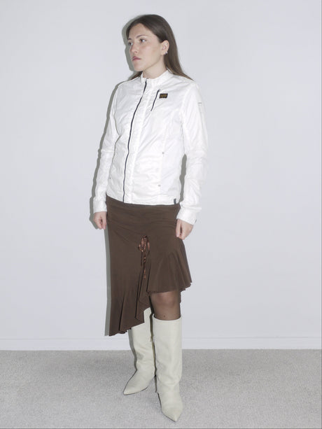 Model wearing a fitted white zip-up jacket with logo patch, a brown asymmetrical jersey skirt with drawstring detail, and off-white cowboy boots. A mix of early 2000s sport and femme — like G-Star meets Roberto Cavalli.