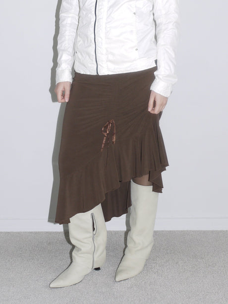 Close-up of a brown asymmetrical jersey skirt with layered hem and ruched satin drawstring detail, styled with off-white vintage cowboy boots. A soft Y2K silhouette with romantic Cavalli energy.