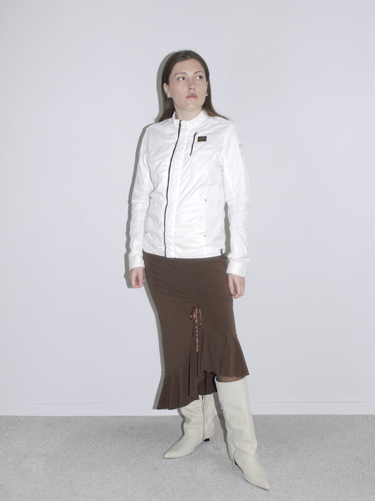 Model wearing a fitted white zip-up jacket with logo patch, a brown asymmetrical jersey skirt with drawstring detail, and off-white cowboy boots. A mix of early 2000s sport and femme — like G-Star meets Roberto Cavalli.