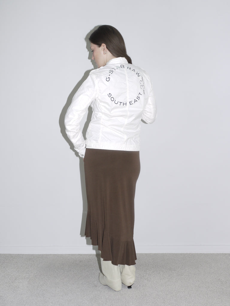 Model wearing a fitted white zip-up jacket with logo patch, a brown asymmetrical jersey skirt with drawstring detail, and off-white cowboy boots. A mix of early 2000s sport and femme — like G-Star meets Roberto Cavalli.
