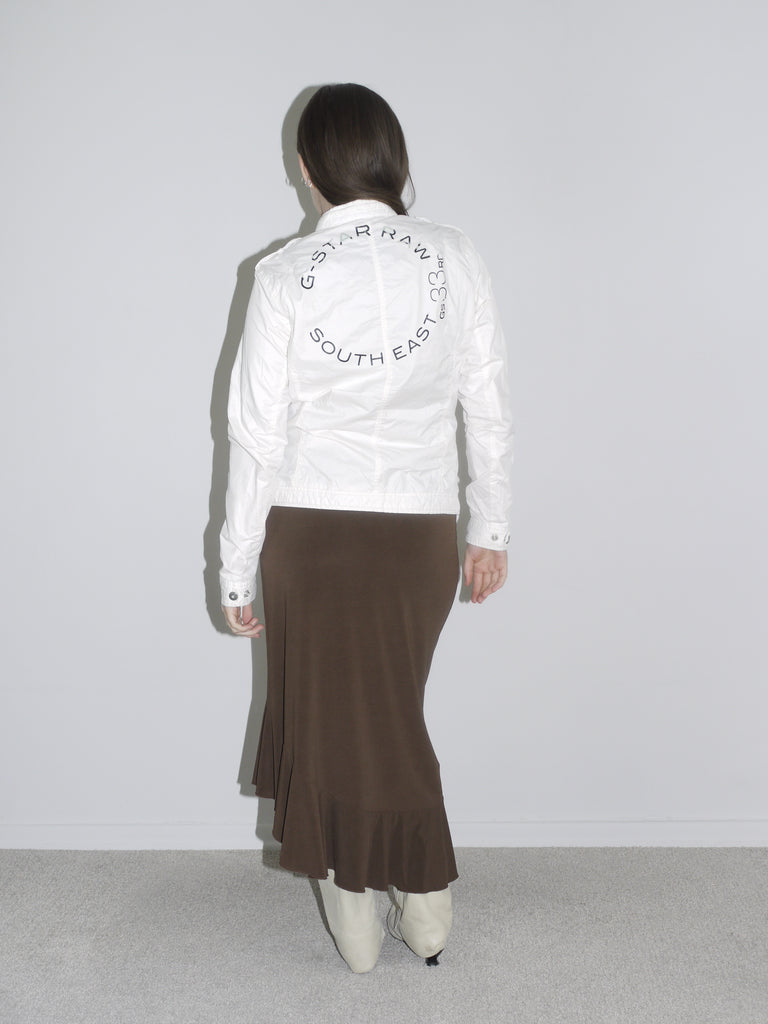 Model wearing a fitted white zip-up jacket with logo patch, a brown asymmetrical jersey skirt with drawstring detail, and off-white cowboy boots. A mix of early 2000s sport and femme — like G-Star meets Roberto Cavalli.