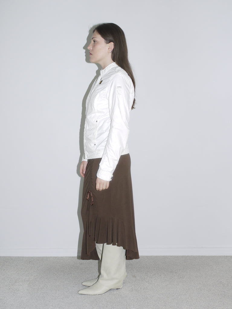 Model wearing a fitted white zip-up jacket with logo patch, a brown asymmetrical jersey skirt with drawstring detail, and off-white cowboy boots. A mix of early 2000s sport and femme — like G-Star meets Roberto Cavalli.