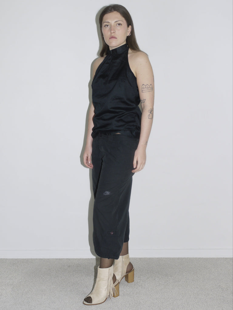 Model wearing a black halterneck top with subtle sheen, paired with cropped Nike cargo pants and beige open-toe fringe heels. A clean yet edgy Y2K silhouette — Margiela meets Nike ACG with a touch of eveningwear.
