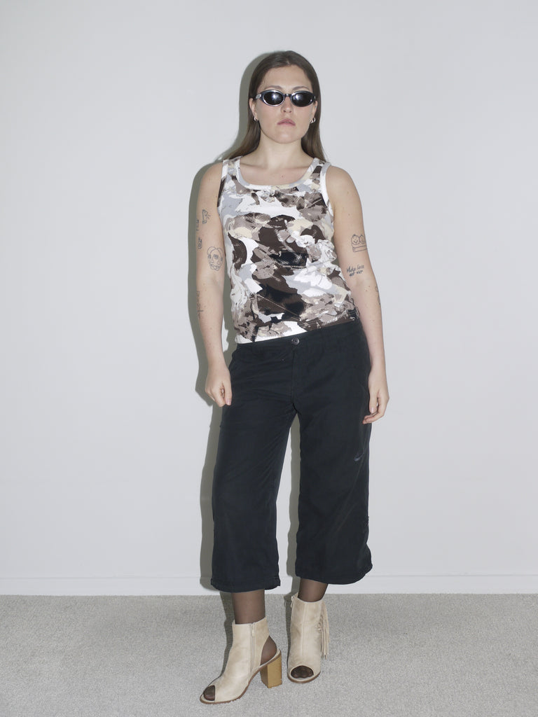 Model wearing a sleeveless abstract-print tank top in earthy tones, paired with low-rise black G-Star inspired trousers and beige open-toe heels. The look is completed with slim black sunglasses and visible tattoos — bold, early-2000s energy with a utilitarian twist.