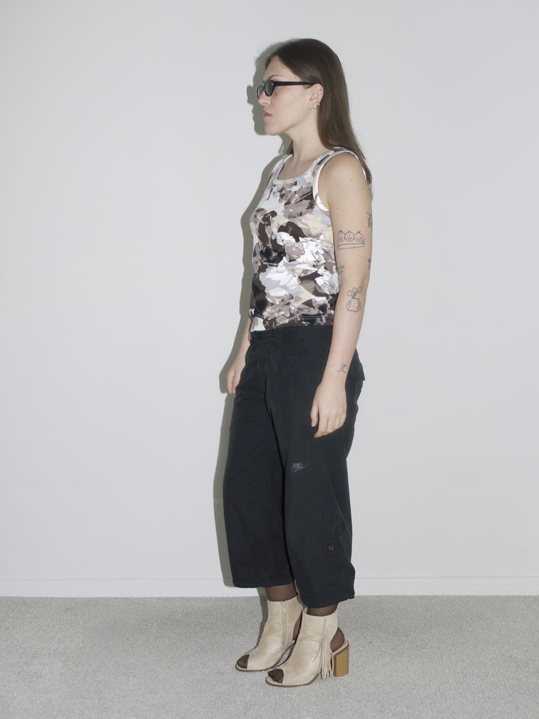Model wearing a sleeveless abstract-print tank top in earthy tones, paired with low-rise black G-Star inspired trousers and beige open-toe heels. The look is completed with slim black sunglasses and visible tattoos — bold, early-2000s energy with a utilitarian twist.