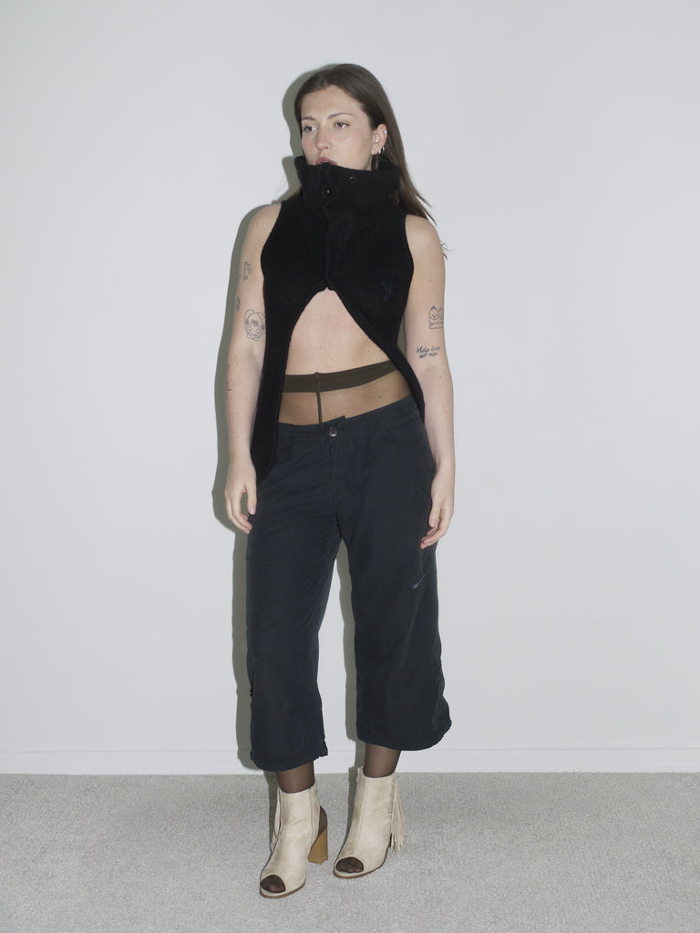 Model in a sculptural black sleeveless zip-up top with high collar and open front, revealing sheer brown tights and low-rise black cargo pants. Styled with beige open-toe boots and visible tattoos — deconstructed elegance with clear G-Star and Margiela-inspired undertones.