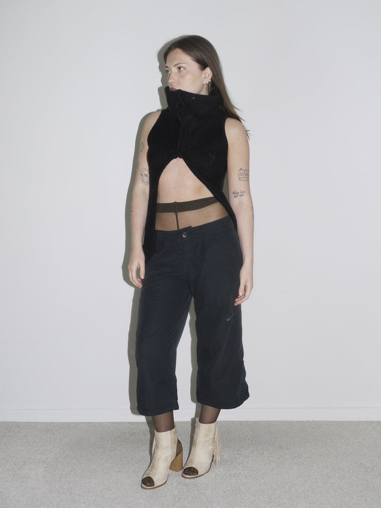 Model in a sculptural black sleeveless zip-up top with high collar and open front, revealing sheer brown tights and low-rise black cargo pants. Styled with beige open-toe boots and visible tattoos — deconstructed elegance with clear G-Star and Margiela-inspired undertones.