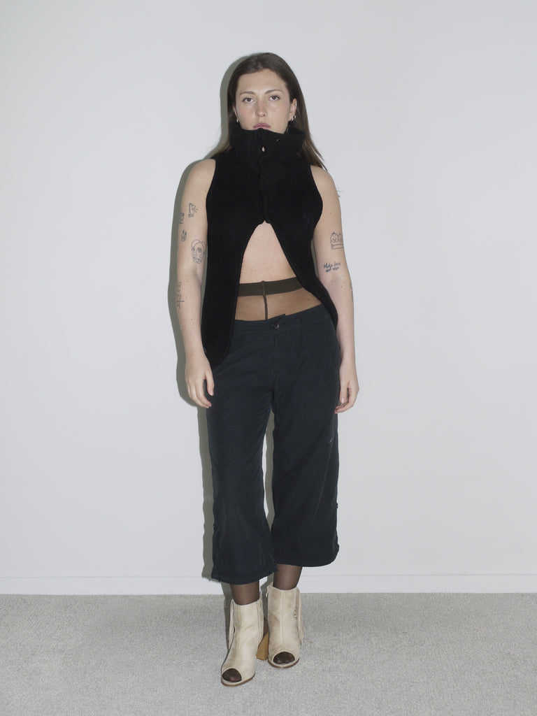 Model in a sculptural black sleeveless zip-up top with high collar and open front, revealing sheer brown tights and low-rise black cargo pants. Styled with beige open-toe boots and visible tattoos — deconstructed elegance with clear G-Star and Margiela-inspired undertones.