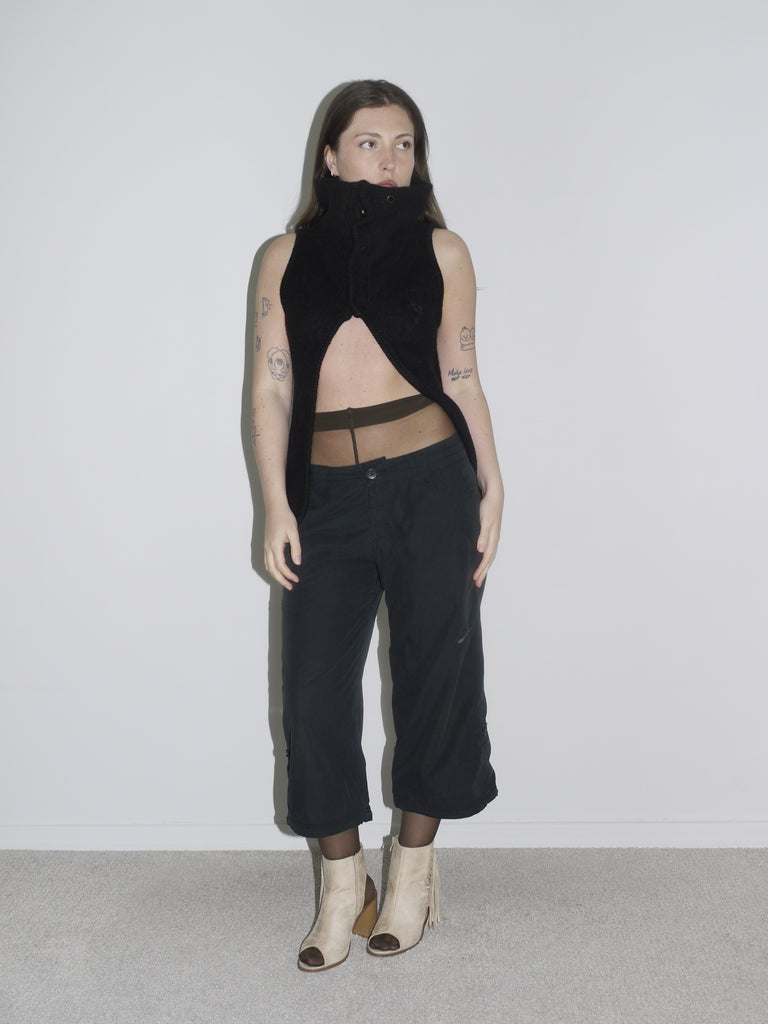 Model in a sculptural black sleeveless zip-up top with high collar and open front, revealing sheer brown tights and low-rise black cargo pants. Styled with beige open-toe boots and visible tattoos — deconstructed elegance with clear G-Star and Margiela-inspired undertones.