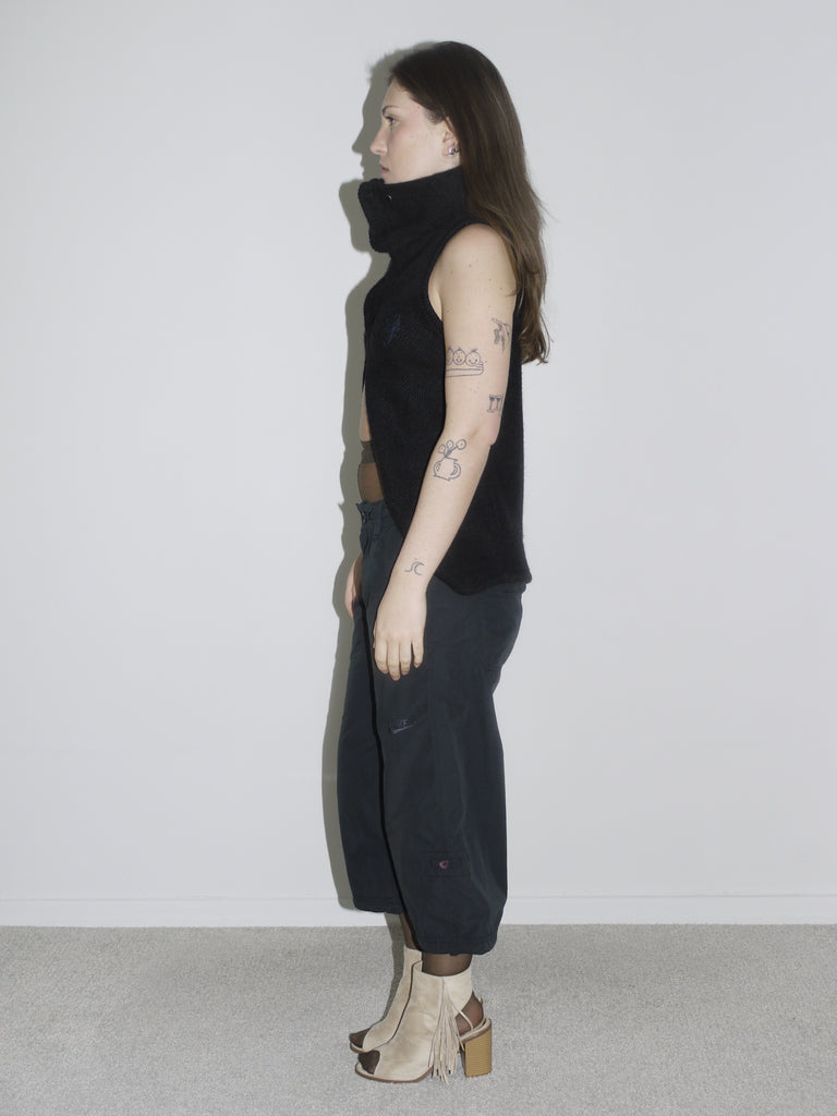 Model in a sculptural black sleeveless zip-up top with high collar and open front, revealing sheer brown tights and low-rise black cargo pants. Styled with beige open-toe boots and visible tattoos — deconstructed elegance with clear G-Star and Margiela-inspired undertones.