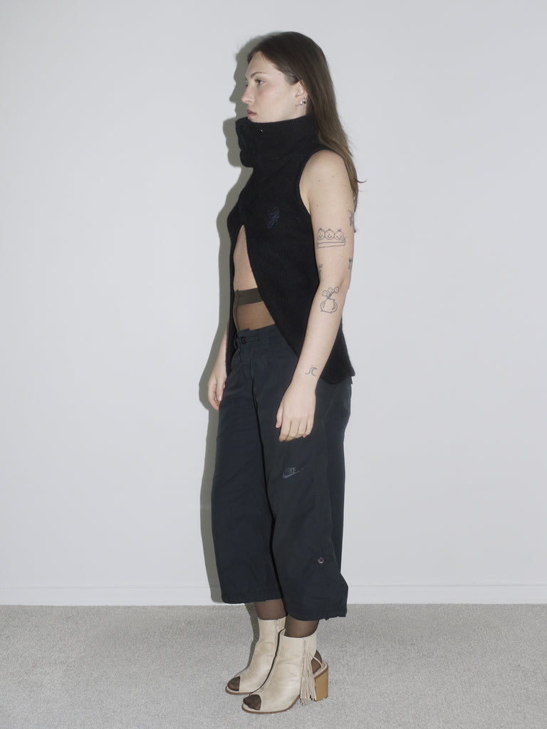 Model in a sculptural black sleeveless zip-up top with high collar and open front, revealing sheer brown tights and low-rise black cargo pants. Styled with beige open-toe boots and visible tattoos — deconstructed elegance with clear G-Star and Margiela-inspired undertones.