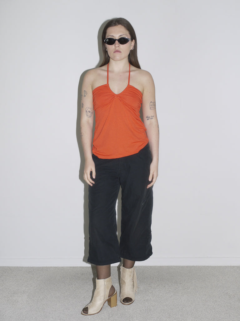 Model in a ruched orange halter top paired with black cropped cargo pants and beige open-toe block heel boots. Styled with sheer tights and slim black sunglasses — early-2000s clubwear meets G-Star utility, bold and minimal at once.