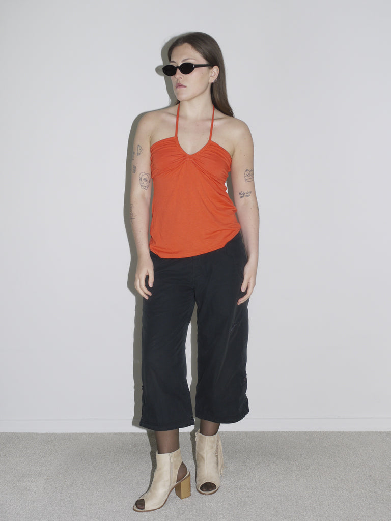 Model in a ruched orange halter top paired with black cropped cargo pants and beige open-toe block heel boots. Styled with sheer tights and slim black sunglasses — early-2000s clubwear meets G-Star utility, bold and minimal at once.