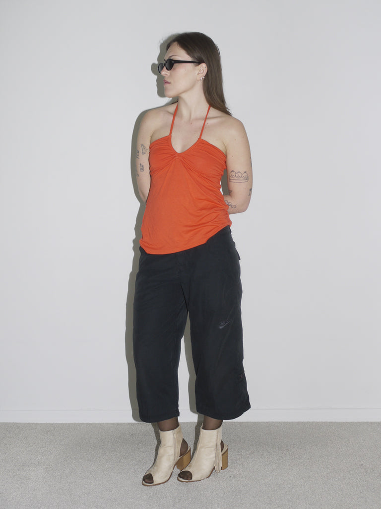Model in a ruched orange halter top paired with black cropped cargo pants and beige open-toe block heel boots. Styled with sheer tights and slim black sunglasses — early-2000s clubwear meets G-Star utility, bold and minimal at once.