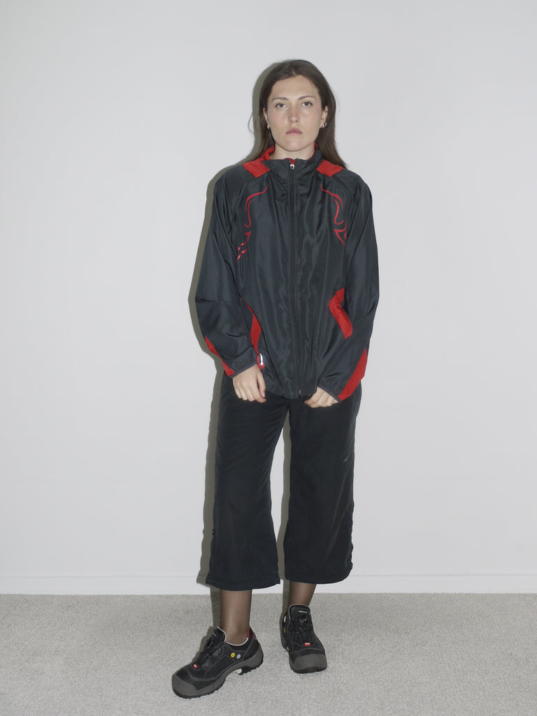 Model wearing a black zip-up windbreaker with red tribal-style racing details, paired with black cropped cargo pants and chunky black sneakers. The look blends Y2K motorsport energy with urban utilitarianism — reminiscent of early 2000s Puma, McQueen x Puma, and underground techwear.
