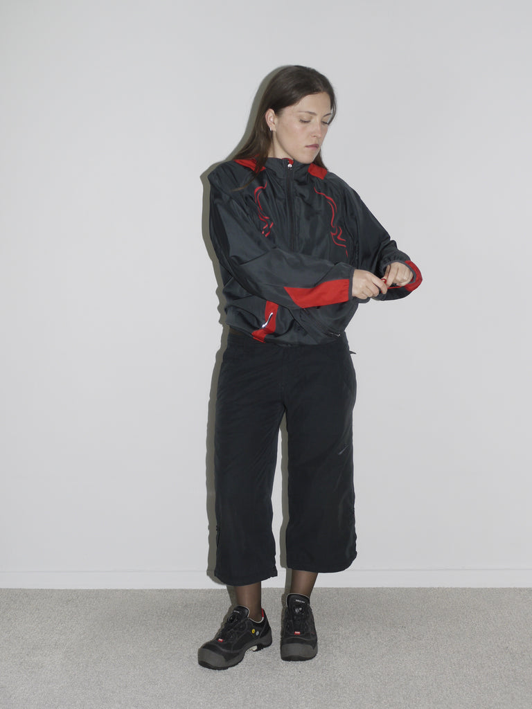 Model wearing a black zip-up windbreaker with red tribal-style racing details, paired with black cropped cargo pants and chunky black sneakers. The look blends Y2K motorsport energy with urban utilitarianism — reminiscent of early 2000s Puma, McQueen x Puma, and underground techwear.