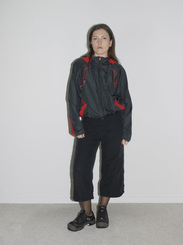 Model wearing a black zip-up windbreaker with red tribal-style racing details, paired with black cropped cargo pants and chunky black sneakers. The look blends Y2K motorsport energy with urban utilitarianism — reminiscent of early 2000s Puma, McQueen x Puma, and underground techwear.