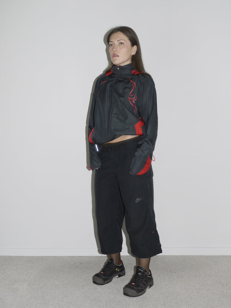 Model wearing a black zip-up windbreaker with red tribal-style racing details, paired with black cropped cargo pants and chunky black sneakers. The look blends Y2K motorsport energy with urban utilitarianism — reminiscent of early 2000s Puma, McQueen x Puma, and underground techwear.