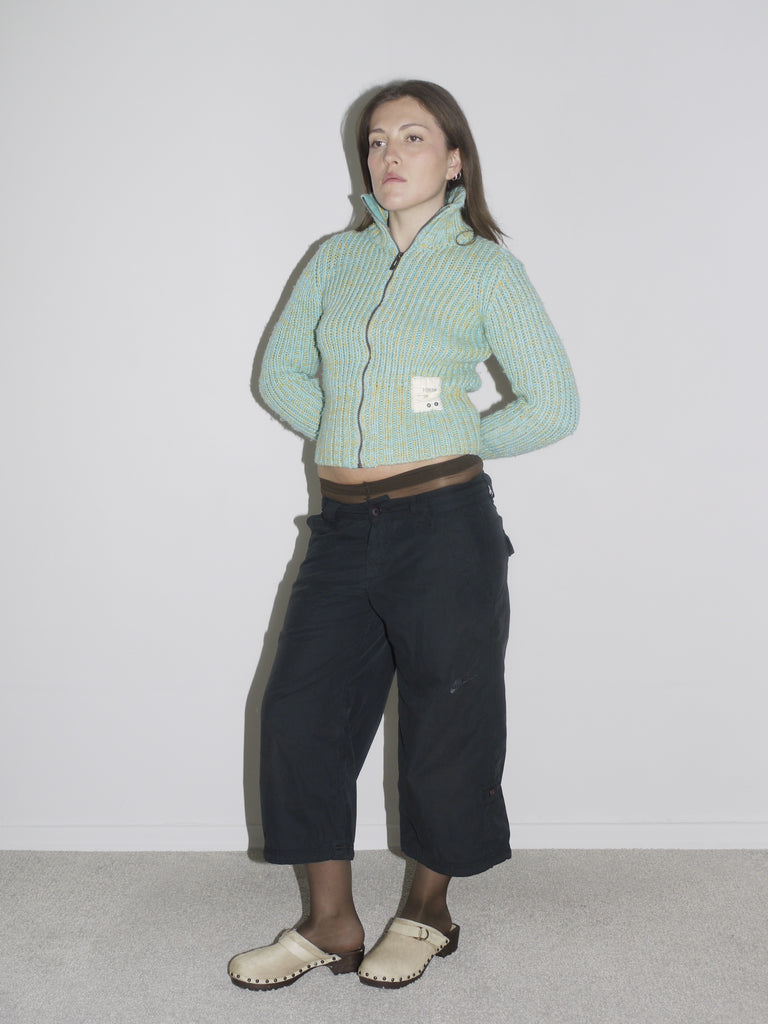 Model wearing a cropped, mint green and yellow ribbed zip-up knit cardigan with a visible label patch, paired with black vintage Nike cargo pants and beige wooden-sole clogs. A playful take on utility wear — think Paloma Wool meets Nike ACG with a hint of Marni.