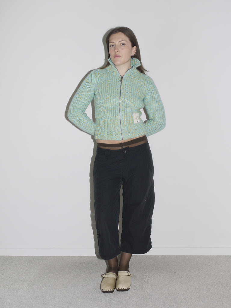 Model wearing a cropped, mint green and yellow ribbed zip-up knit cardigan with a visible label patch, paired with black vintage Nike cargo pants and beige wooden-sole clogs. A playful take on utility wear — think Paloma Wool meets Nike ACG with a hint of Marni.
