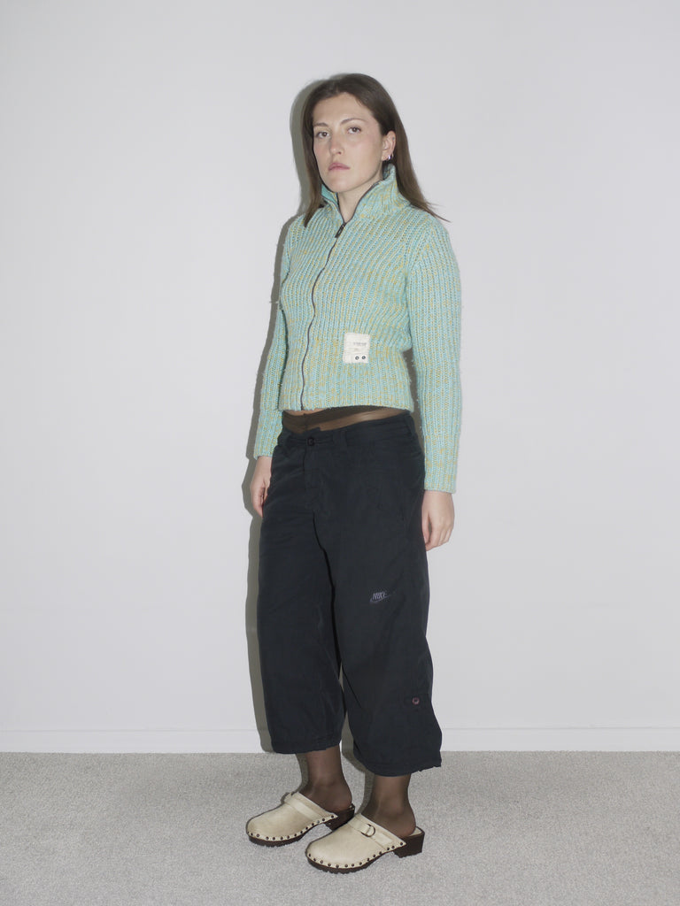 Model wearing a cropped, mint green and yellow ribbed zip-up knit cardigan with a visible label patch, paired with black vintage Nike cargo pants and beige wooden-sole clogs. A playful take on utility wear — think Paloma Wool meets Nike ACG with a hint of Marni.