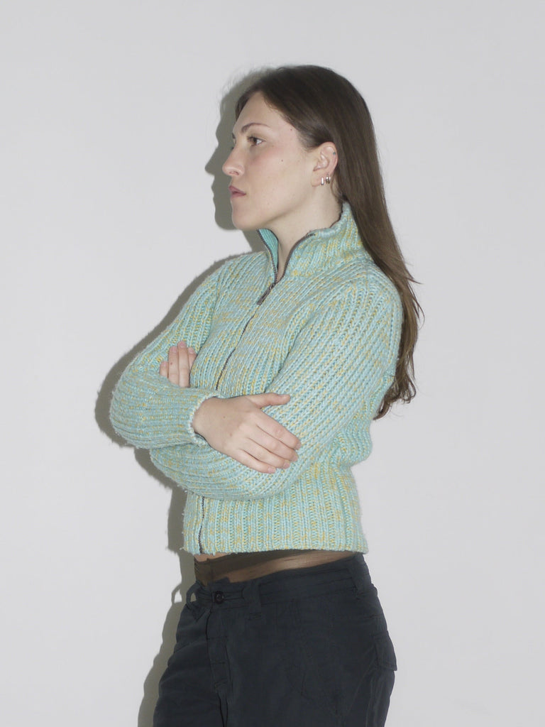 Model wearing a cropped, mint green and yellow ribbed zip-up knit cardigan with a visible label patch, paired with black vintage Nike cargo pants and beige wooden-sole clogs. A playful take on utility wear — think Paloma Wool meets Nike ACG with a hint of Marni.