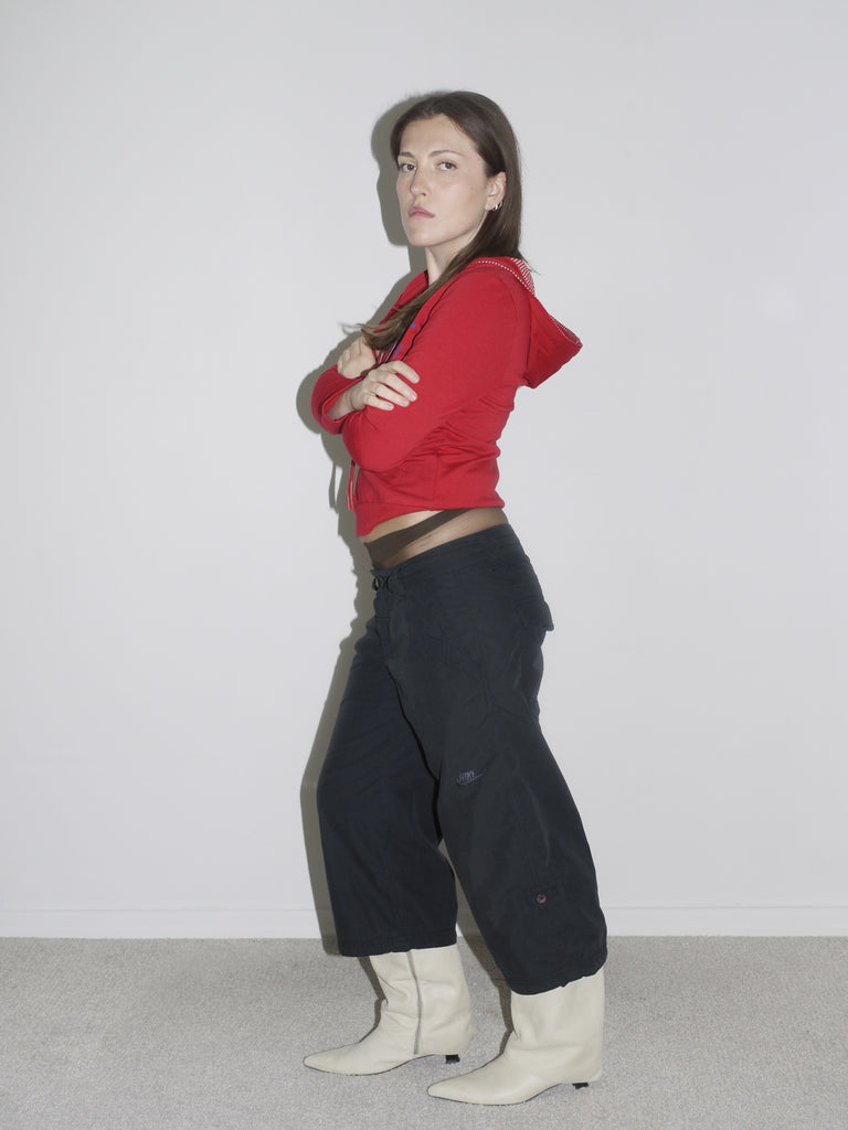 Model wearing a fitted red zip-up hoodie with studded hood detail, low-rise black Nike cargo pants, and pointed white leather cowboy boots. The look captures peak Y2K attitude — part Britney, part G-Star, part Western revival.